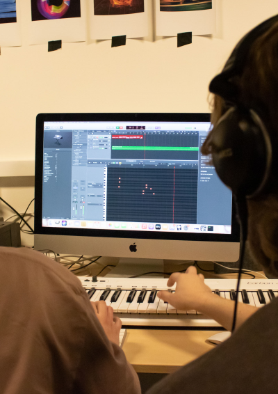 To personer med høretelefoner sidder omkring en iMac og producerer musik. Den ene spiller på et midi-keyboard.